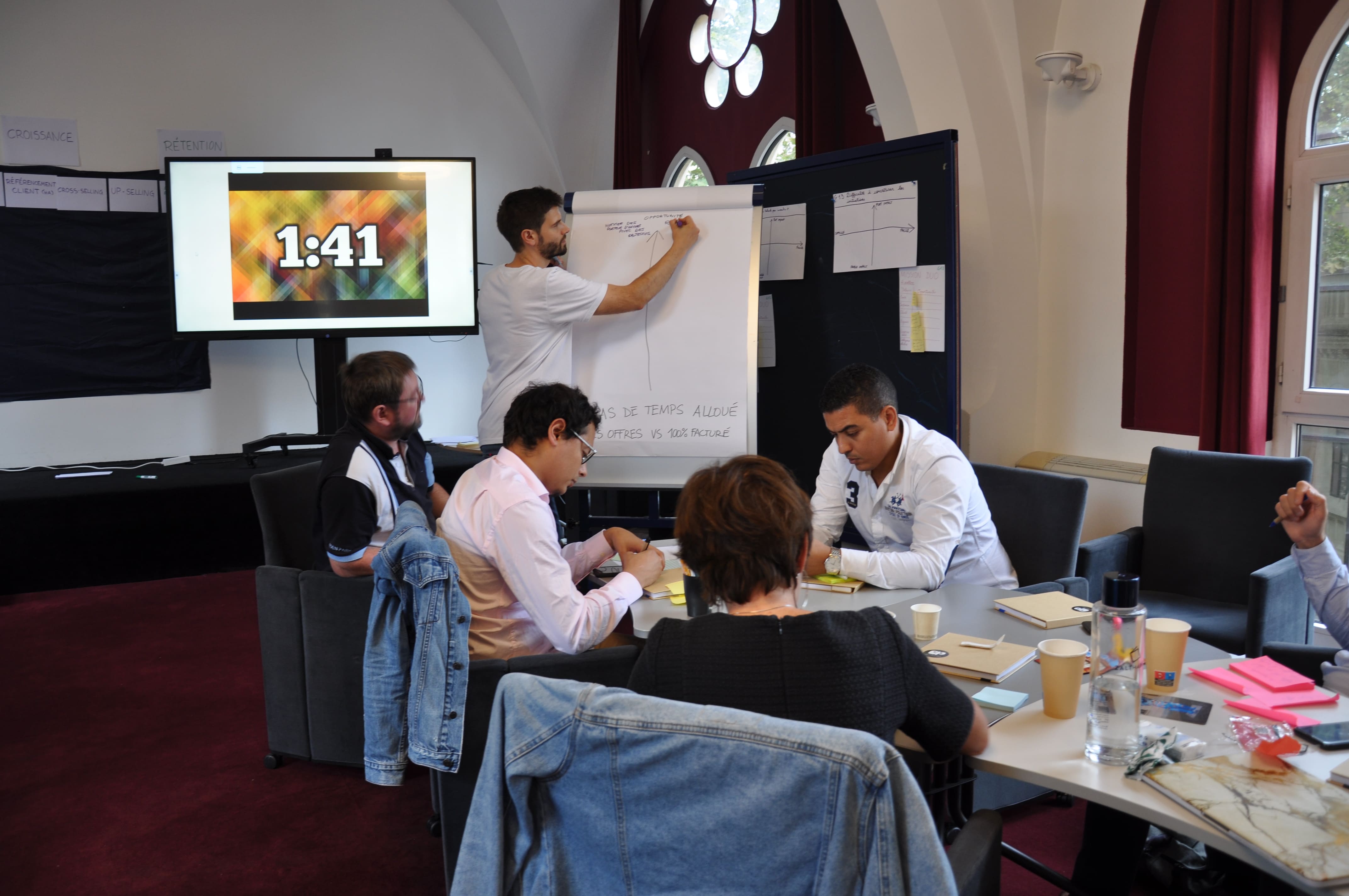 Quentin écrit au paperboard pendant qu'un timer 1:41 défile sur l'écran à côté. Plusieurs participants prennent des notes assis à la table.