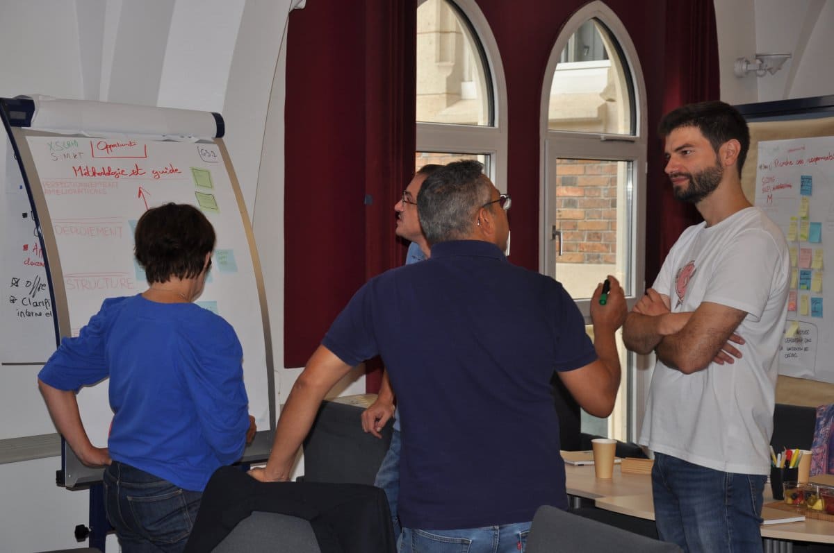 Quentin en T-shirt blanc anime un atelier face à un paperboard listant les axes : Méthodologie et guide, Déploiement, Structure, Offre. Trois participants discutent autour de lui dans une salle aux fenêtres en ogive.